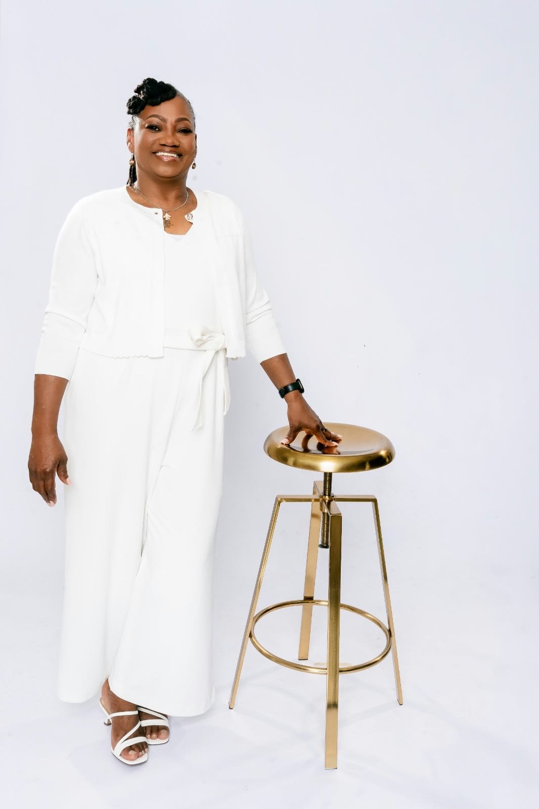 A smiling woman in white standing beside a gold stool.