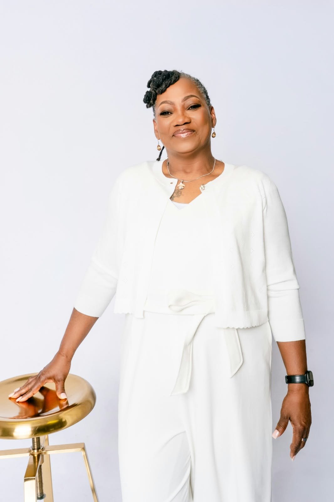 A woman in white resting one hand on a gold stool.